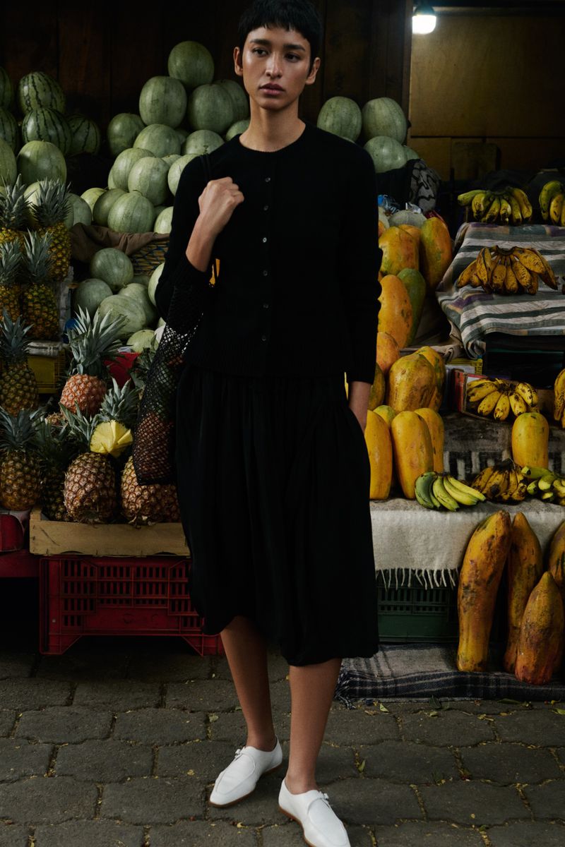 Young adult woman in black dress stands in an outdoor market surrounded by fresh produce in daytime portrait shot.