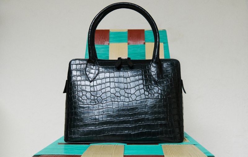 Black alligator-patterned handbag with round handles sits on a colorful woven chair in a studio setting with a pale background.