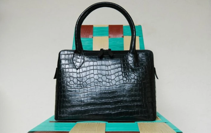 Black alligator-patterned handbag with round handles sits on a colorful woven chair in a studio setting with a pale background.