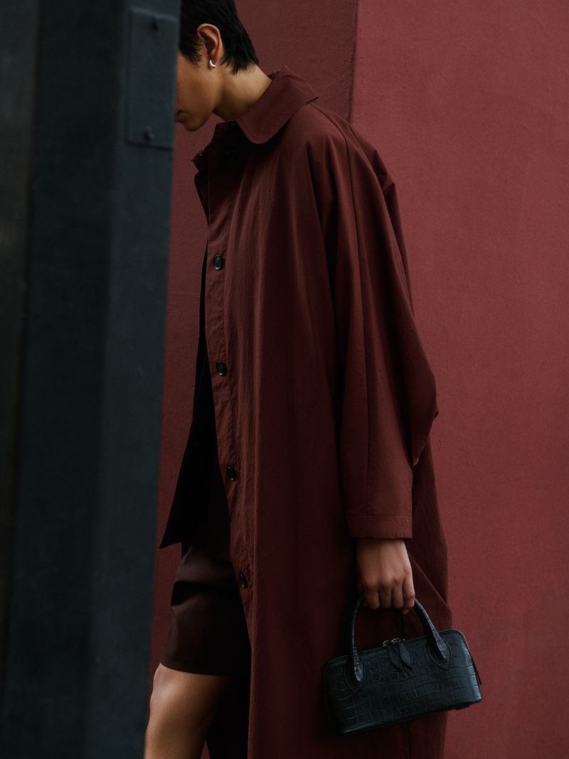 Woman in a brown coat walks past a wall carrying a small, structured black purse in an outdoor, urban environment.