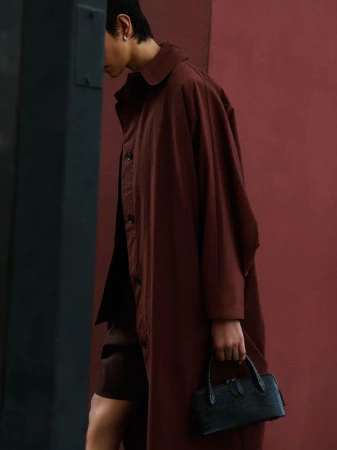 Woman in a brown coat walks past a wall carrying a small, structured black purse in an outdoor, urban environment.