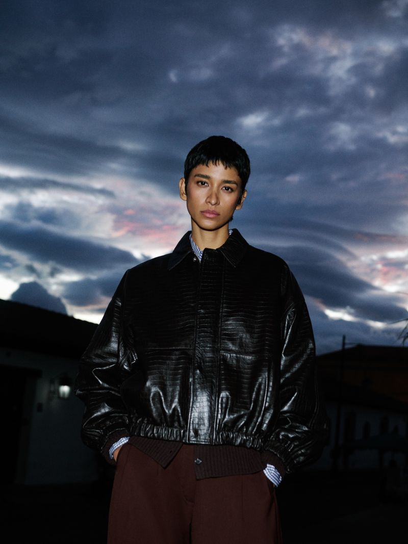 A young woman with a short hairstyle poses outdoors at dusk, wearing a black leather-look jacket, brown pants, and patterned shirt, set against a cloudy sky.