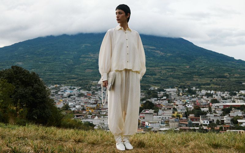 Adult female in a cream-colored, loose-fitting outfit with a purse, standing in a field with a town and mountain in the background.