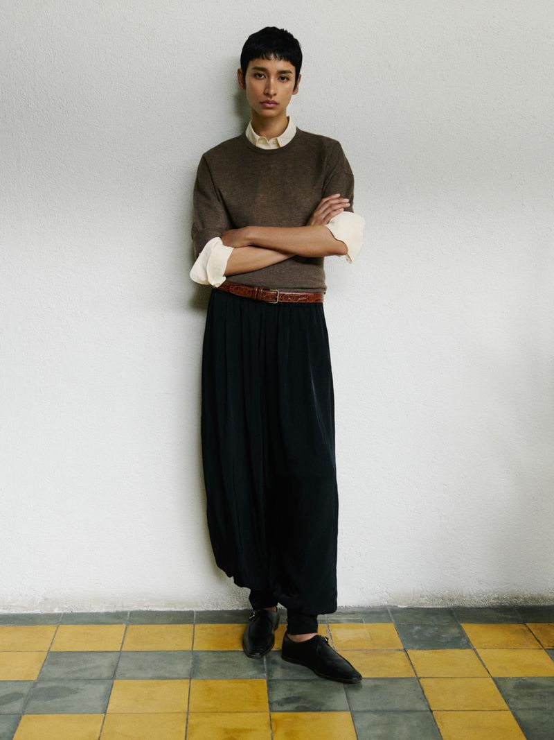 Full-length portrait of an adult female with short dark hair, wearing a brown sweater, black skirt, and black shoes, posing indoors against a white wall.