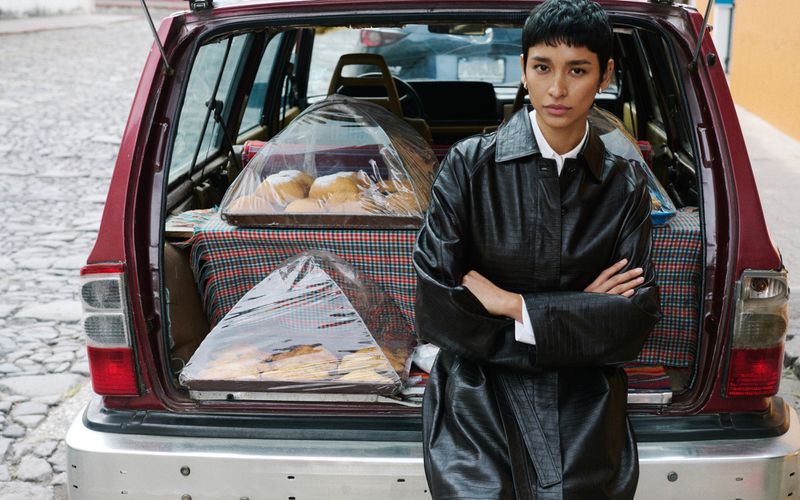 A stylish adult woman poses in front of the open trunk of a maroon vehicle displaying trays of pastries on a cobblestone street.