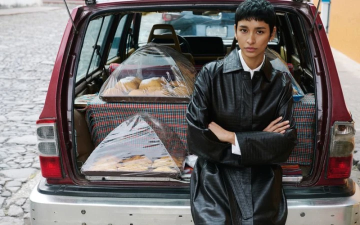 A stylish adult woman poses in front of the open trunk of a maroon vehicle displaying trays of pastries on a cobblestone street.