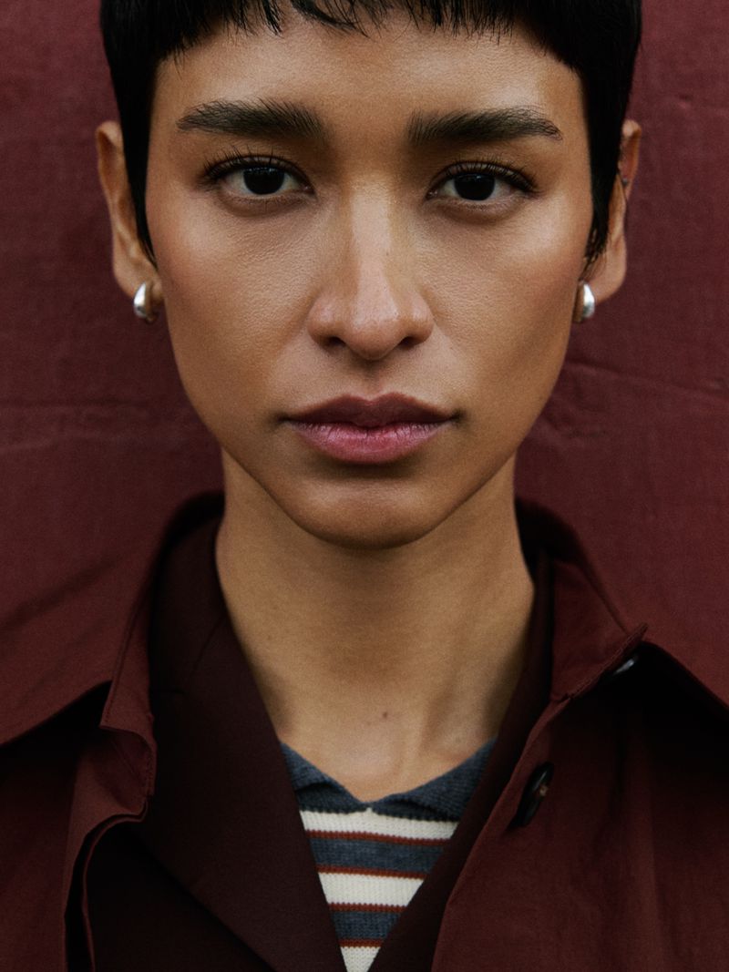 Close-up portrait of a woman with short dark hair, wearing a coat and earrings, against a dark red textured background.