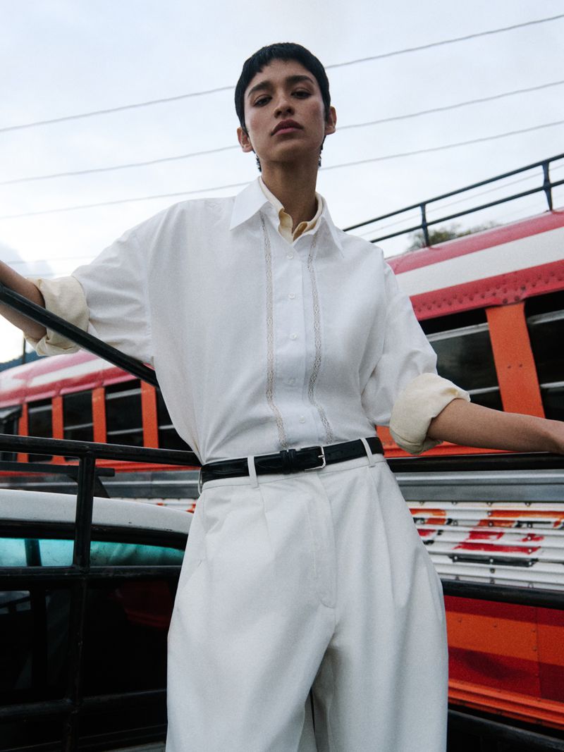 Model Marsella Rea posing in a white button-down shirt and matching pleated trousers beside a red bus for Cordera.