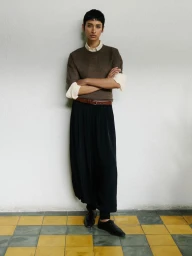 Model standing with arms crossed wearing a brown knit sweater, white collared shirt, and black skirt against a pale wall.