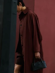 Model carrying a black textured handbag while wearing a long brown overcoat in profile against a dark reddish wall.