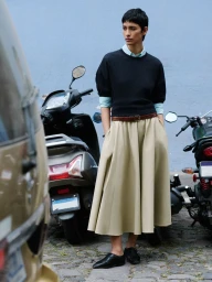 Model Marsella Rea wearing a black sweater and pleated beige midi skirt standing near parked scooters on a street.