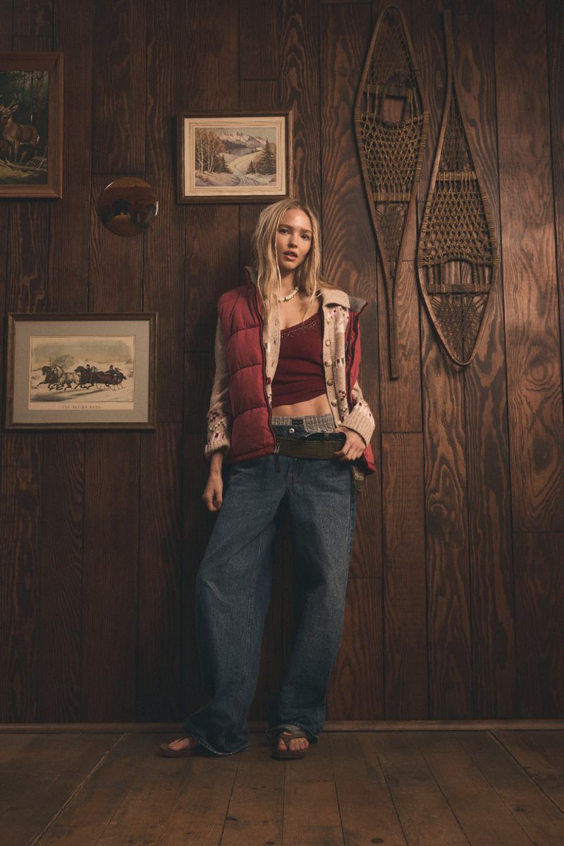 A full-length shot features a blonde woman in a red vest and jeans, posing indoors against a wood-paneled wall decorated with vintage art.