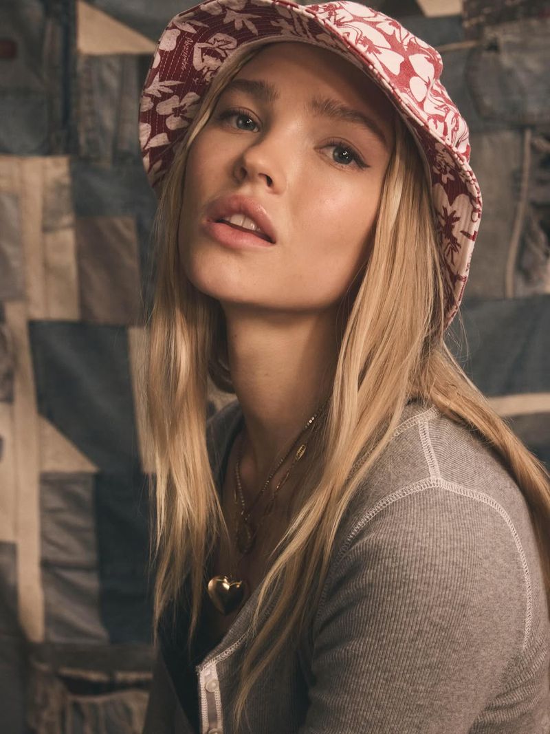 Close-up of a blonde woman wearing a floral bucket hat, gray long-sleeve shirt, and gold necklace with a heart pendant, posing indoors.