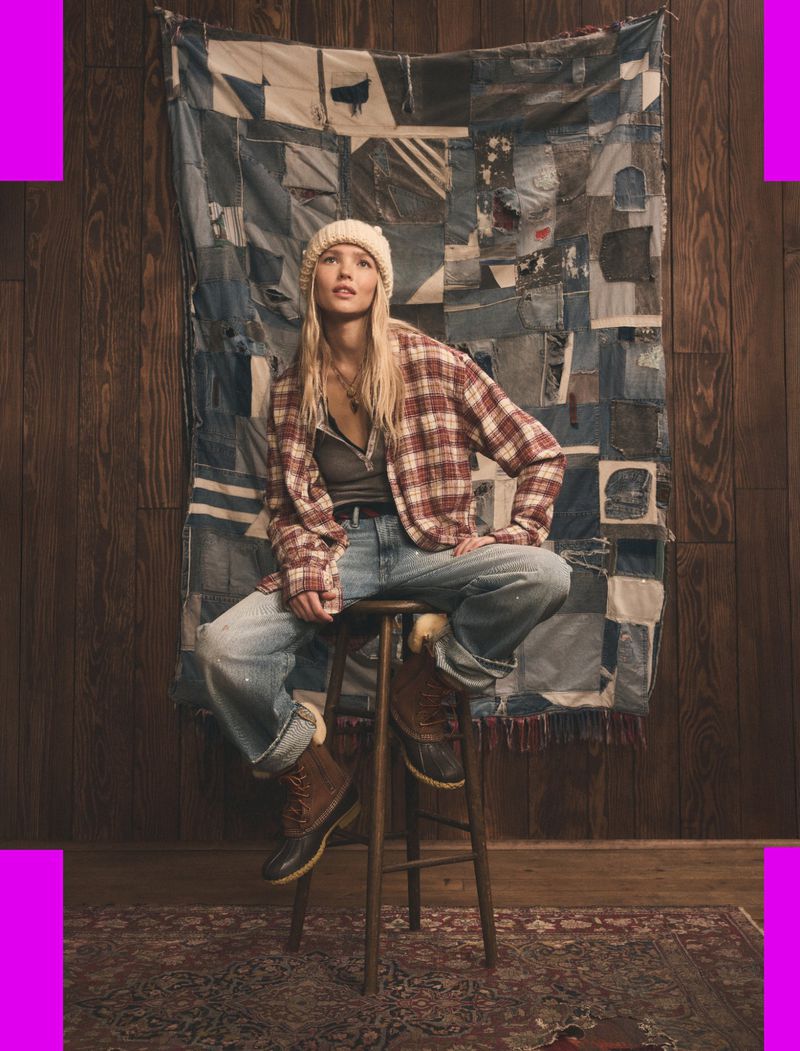 A woman in a knit hat and flannel shirt sits on a stool in a rustic setting with a quilt backdrop, wearing jeans and boots.
