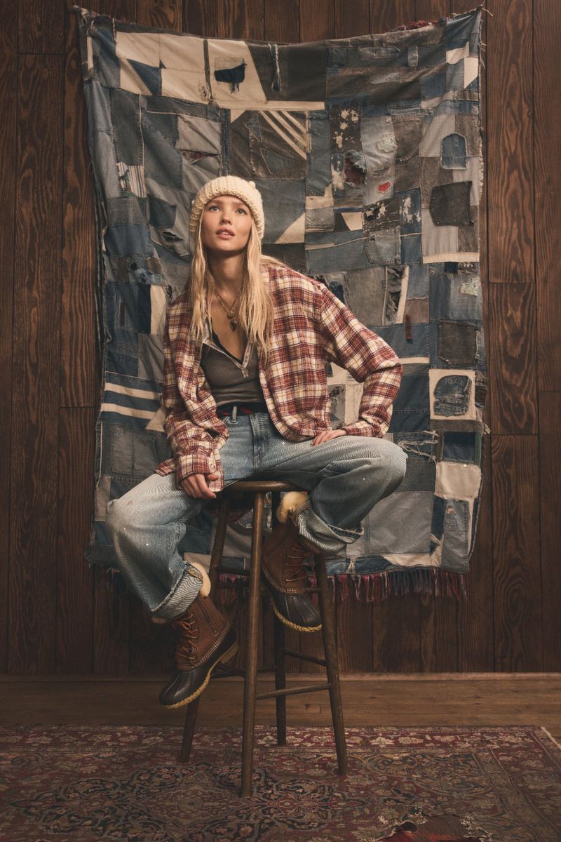 Blonde woman in flannel shirt and jeans sits on a wooden stool in front of a denim patchwork quilt, wearing a beanie.