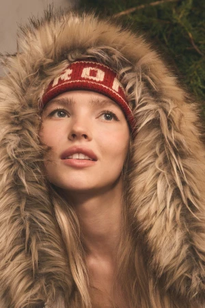 Close-up of a woman with blonde hair wearing a red and white knit headband and fur-trimmed hood. Looking up and to the left.