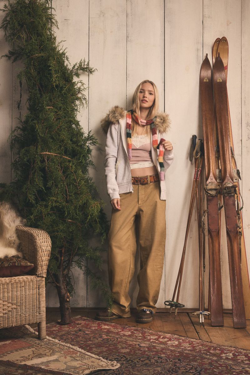 Blonde woman in casual winter fashion, wearing a fur-lined hoodie and khaki pants, poses indoors with a potted tree and skis.