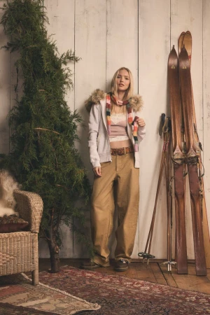 Blonde woman in casual winter fashion, wearing a fur-lined hoodie and khaki pants, poses indoors with a potted tree and skis.