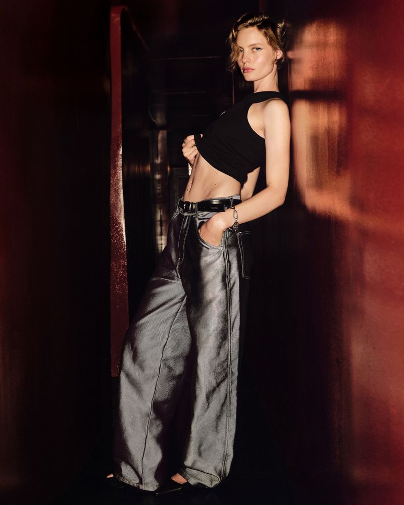 Maya Vozhdaeva poses in a black cropped tank top and silver metallic wide-leg trousers against a dark wall for Calvin Klein.