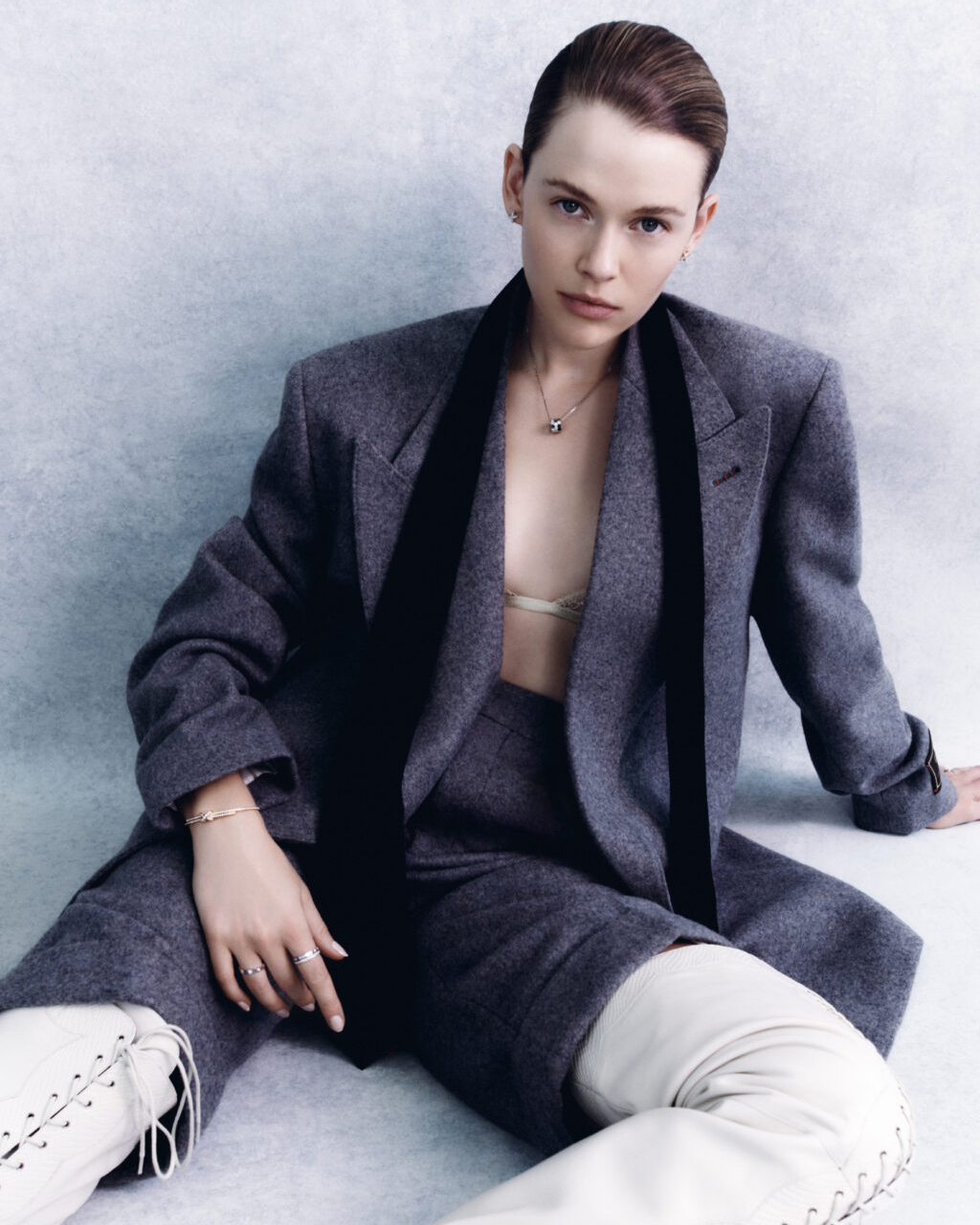 Portrait of a young adult woman in a grey suit jacket and shorts with ivory thigh-high boots sitting against a neutral backdrop.