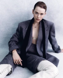 A woman in a grey tailored coat and white lace-up boots sitting against a mottled light blue studio backdrop.