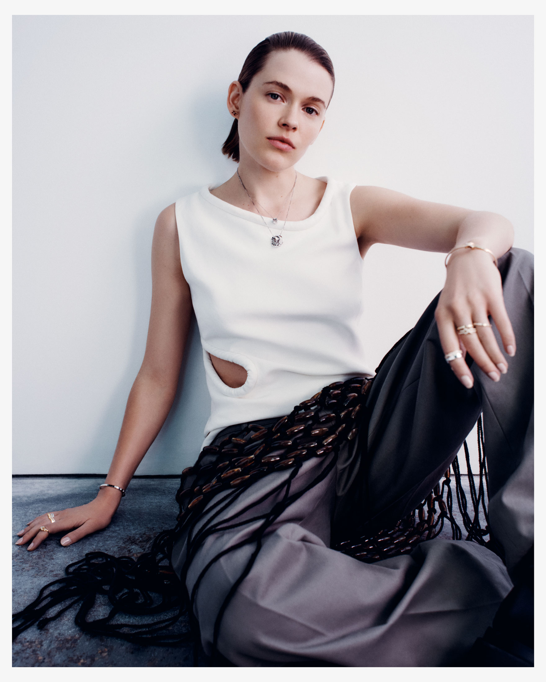 Indoor shot of a woman in casual clothing and jewelry sitting against a white wall, styled with dark pants and top with a cutout.