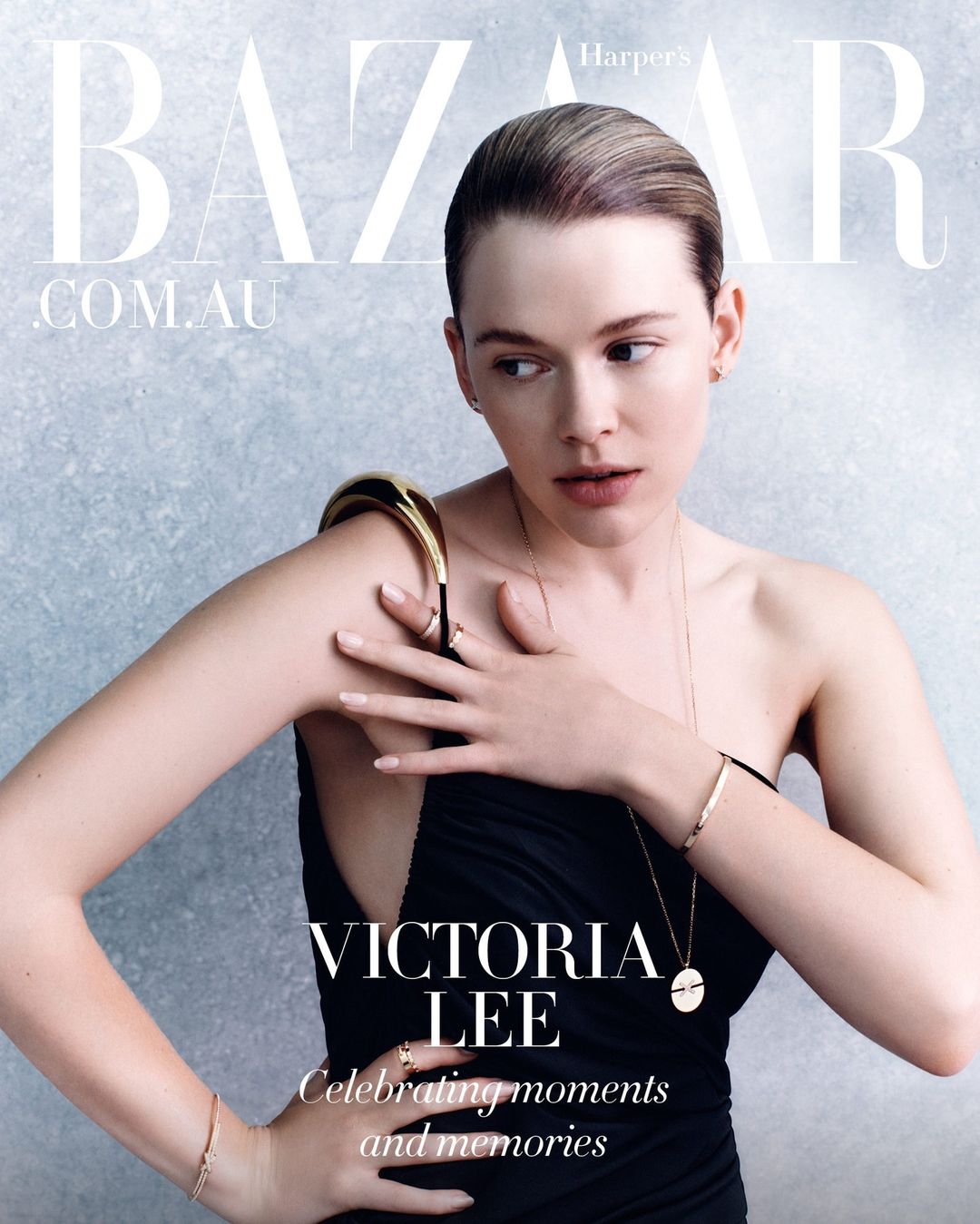 Model Victoria Lee wearing a black dress and gold jewelry against a textured grey background for Harper's Bazaar Australia.