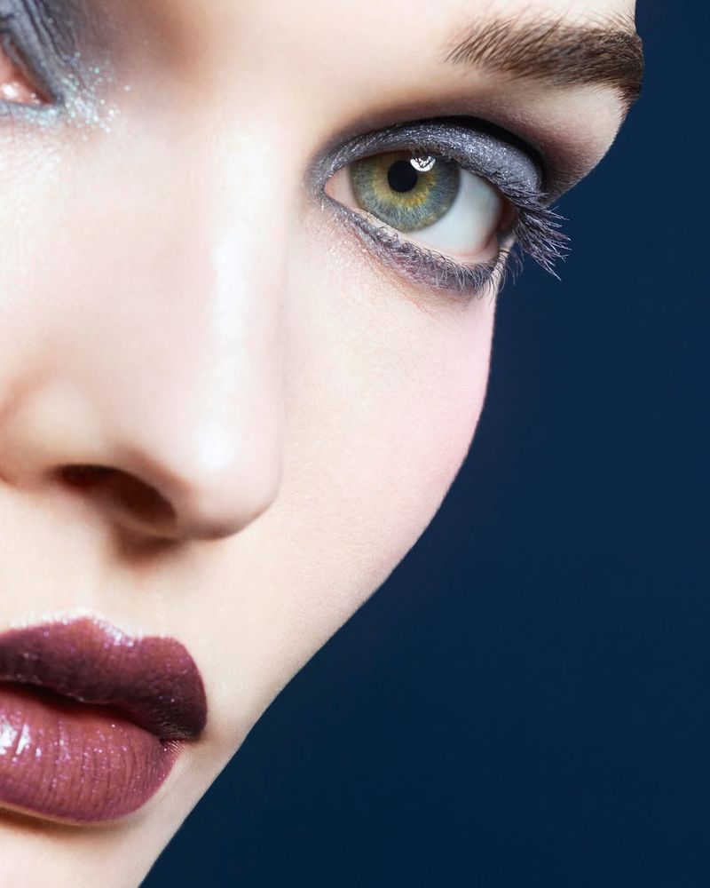 Close-up of a woman's face with dramatic makeup, featuring heavily shadowed eyeshadow and glossy lips against a deep blue background.