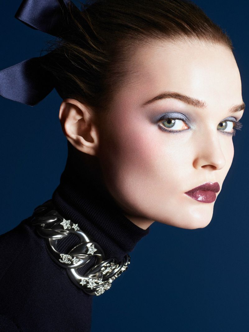 Close-up portrait of a woman with dark hair and makeup, wearing a black turtleneck sweater and a silver chain necklace on a dark blue background.