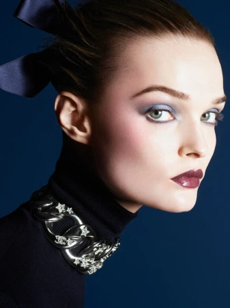 Close-up portrait of a woman with dark hair and makeup, wearing a black turtleneck sweater and a silver chain necklace on a dark blue background.