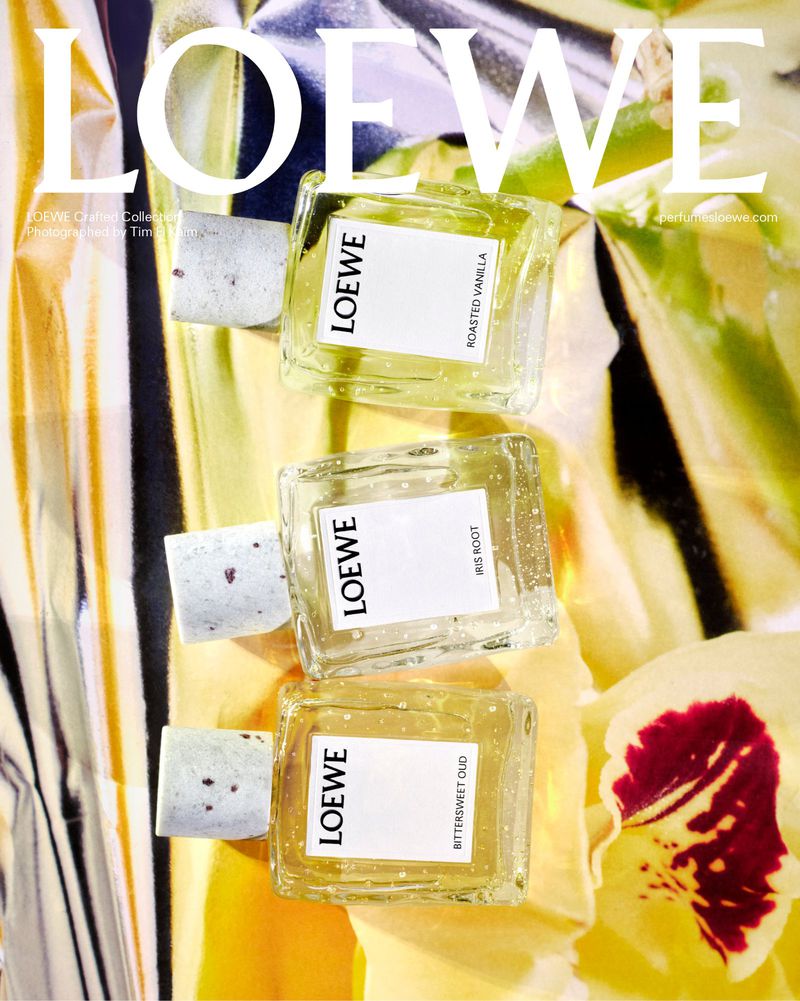 Three LOEWE perfume bottles, each with unique scent labels, are displayed against a backdrop of a shiny gold fabric and white flowers.