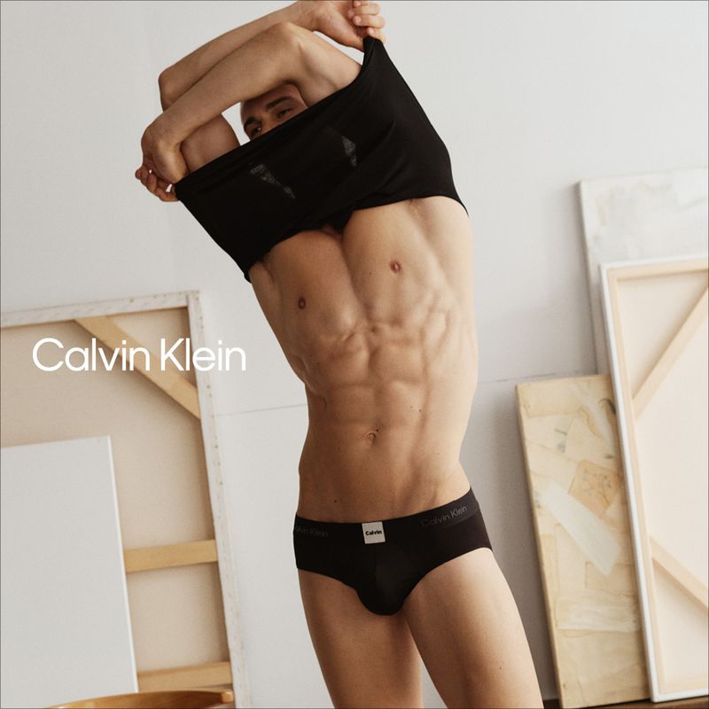 Adult male model posing in black briefs and taking off a black t-shirt inside a photo studio with white walls and canvas.