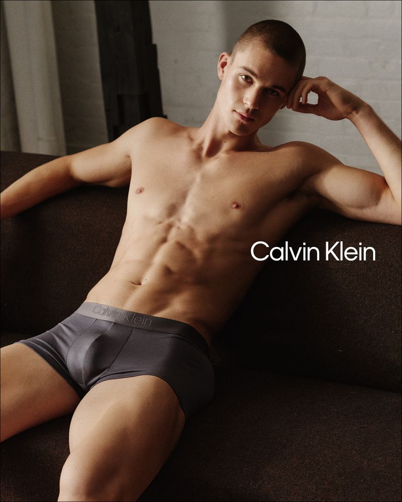 Male underwear model poses on a brown couch in a studio setting. Abs visible. Calvin Klein logo is visible in the background.