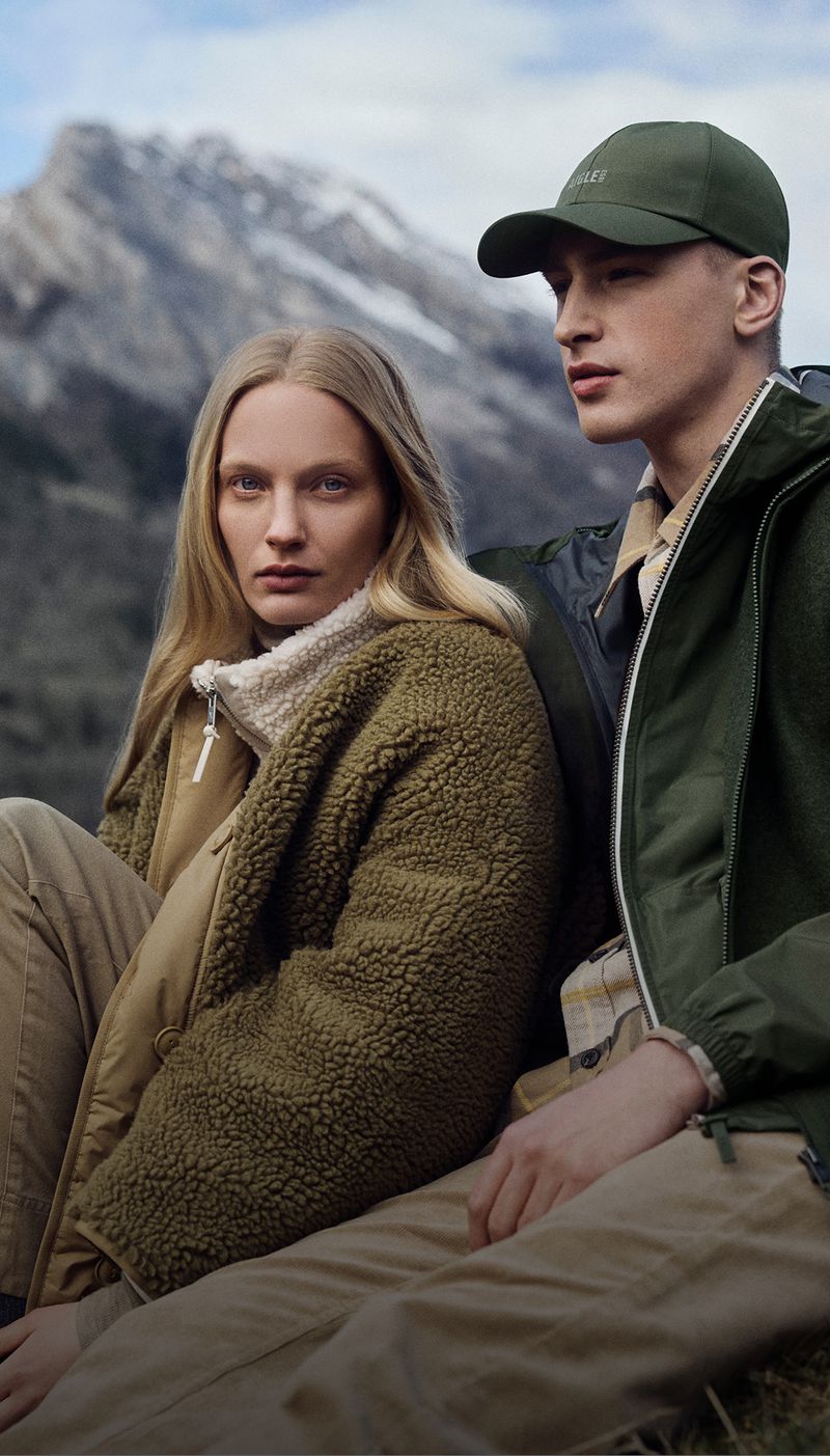 Outdoor fashion shot: a young adult man and woman in jackets and hats, sitting in a field with mountains in the background.