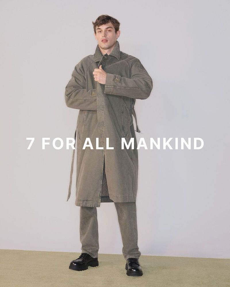 Man poses in long olive-green coat and matching pants against a neutral backdrop, showcasing fashion with a modern, minimalist aesthetic.