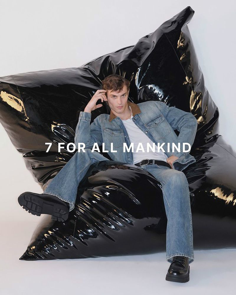 An adult male poses on a large, shiny black beanbag chair in a studio wearing a denim jacket and jeans with black boots, against a white backdrop.