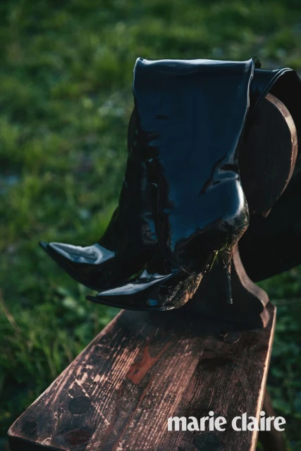 Black patent leather thigh high heel boots on a dark wood bench in outdoor setting with lush green grass backdrop.