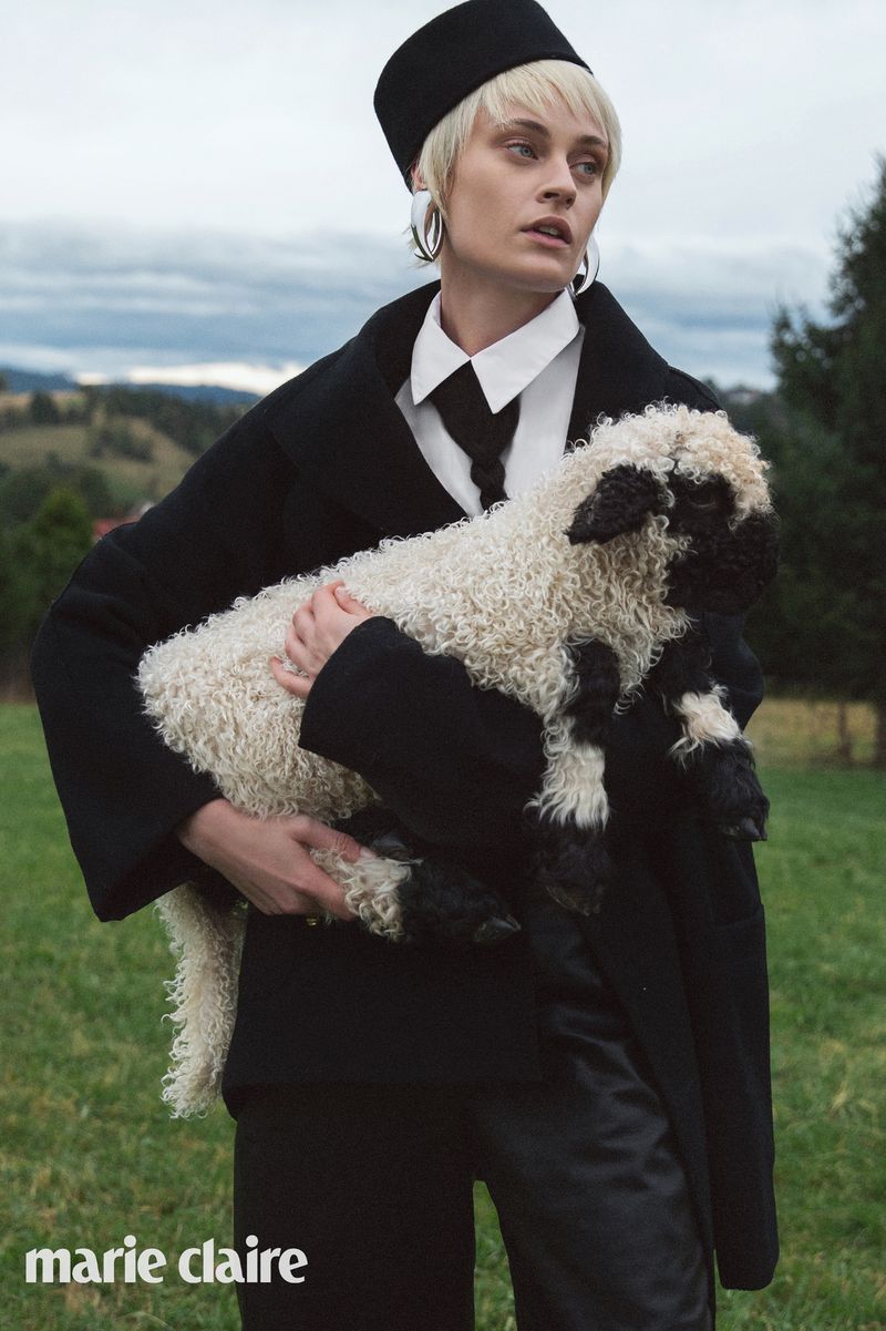 A young woman in a black outfit and hat holds a lamb in a grassy field beneath a cloudy sky, editorial fashion, Marie Claire logo.