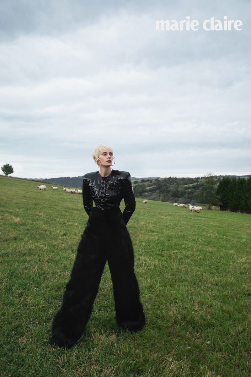 Model Paulina Heiler posing in an all-black structured outfit and oversized wide-leg trousers in an open grassy pasture with sheep.