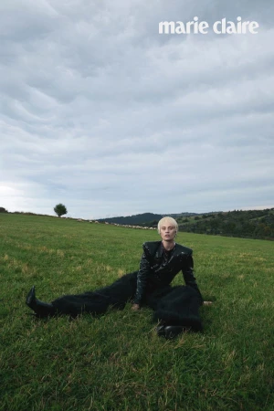 Model Paulina Heiler wearing an all-black structured outfit and boots, reclining on a grassy hill under an overcast sky.