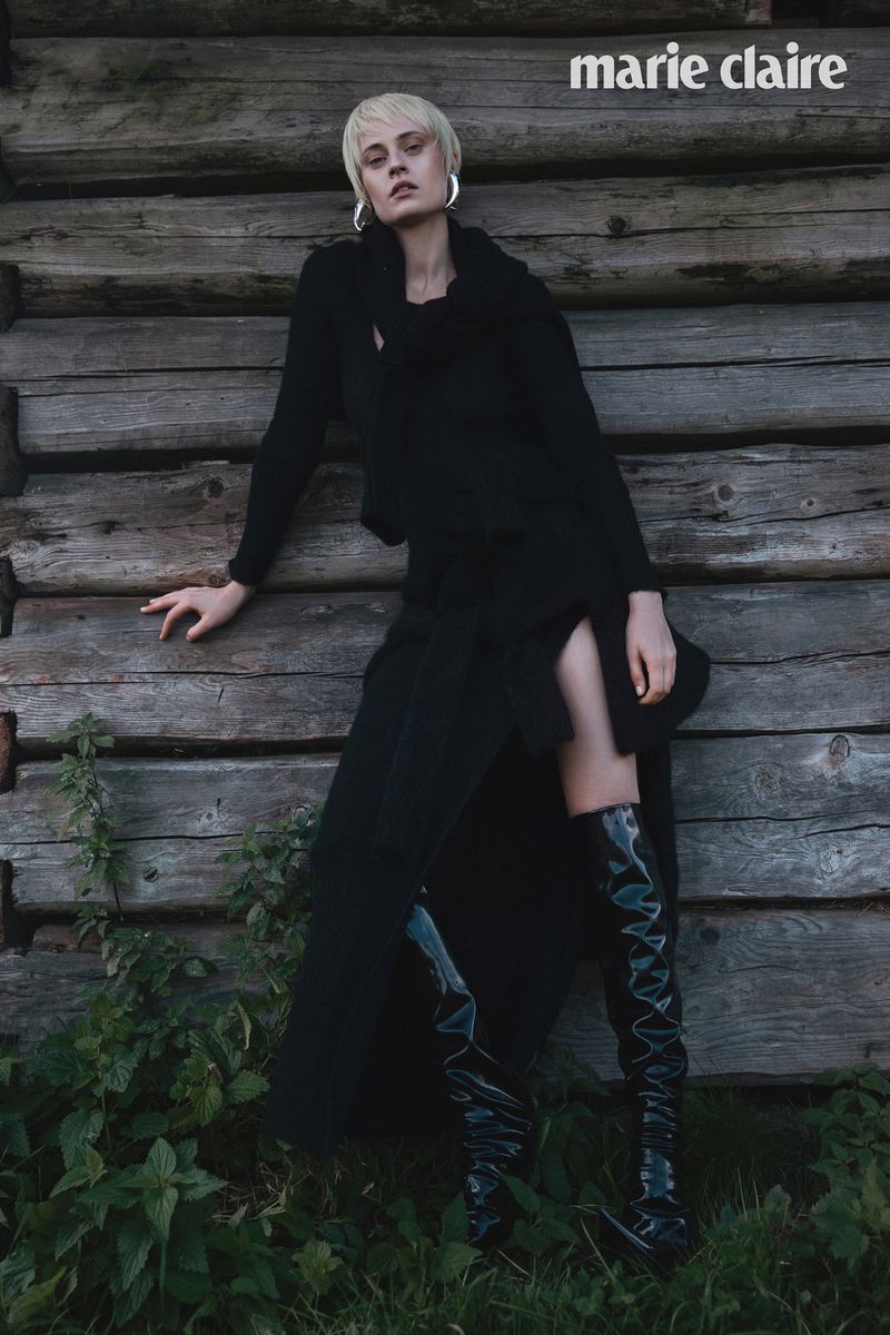 Fashion shot of a blond woman in a black dress and boots posing against a weathered wooden wall with green plants at the bottom.