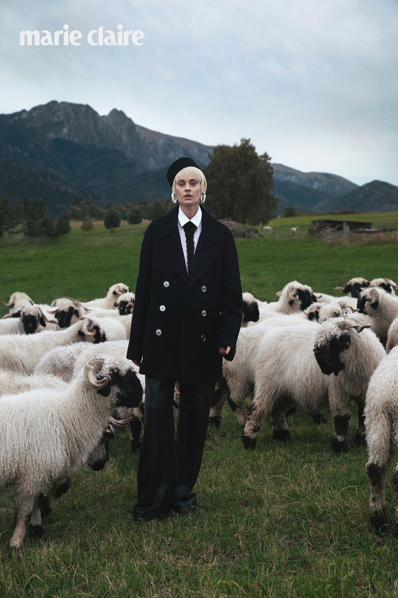 Model Paulina Heiler in a black double-breasted coat and tie standing amidst a flock of sheep in a mountainous meadow.
