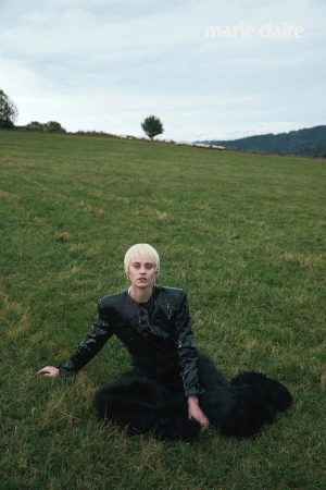Model with blonde hair wearing a black vinyl jacket seated on a lush green meadow under an overcast sky.