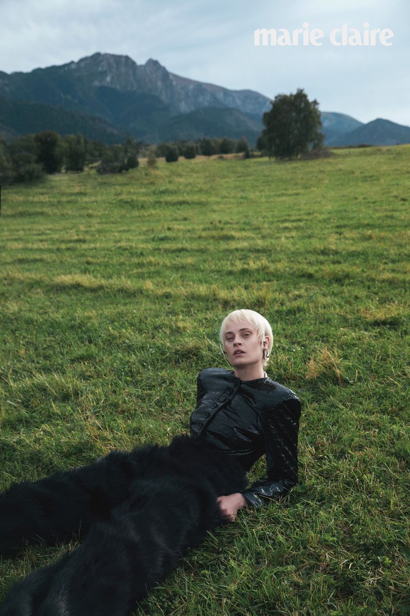 Fashion editorial shot of a woman with short blonde hair wearing a shiny black jacket and fur pants, lying on a grassy field with mountains.