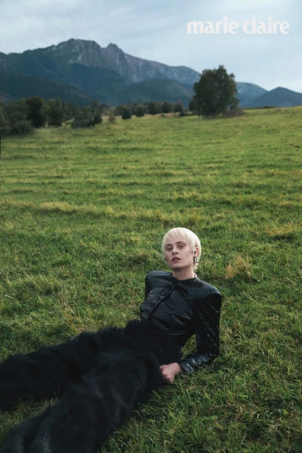 Fashion editorial shot of a woman with short blonde hair wearing a shiny black jacket and fur pants, lying on a grassy field with mountains.