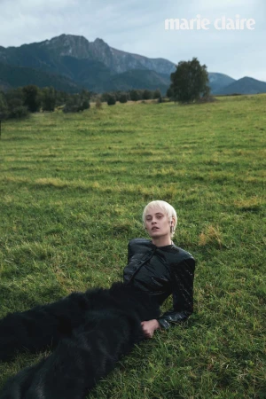 Fashion editorial shot of a woman with short blonde hair wearing a shiny black jacket and fur pants, lying on a grassy field with mountains.