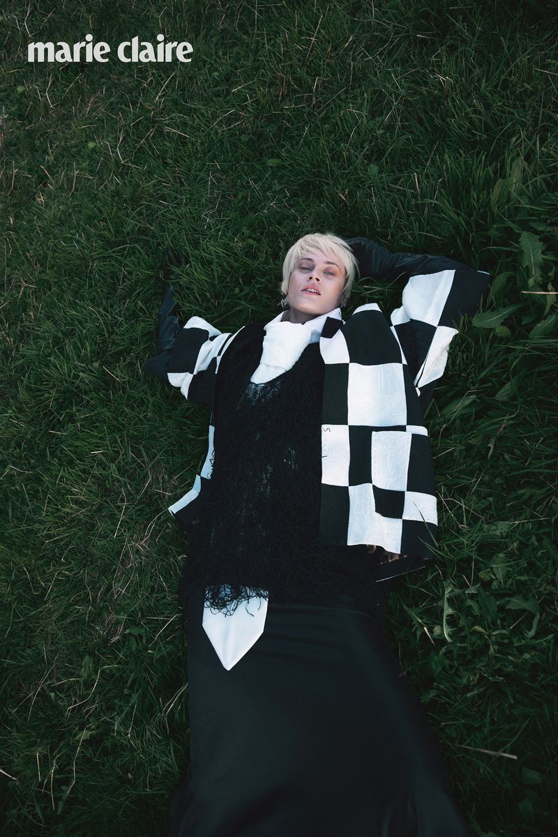 Model lying on deep green grass wearing a black and white checkered jacket with a high white collar and black gloves.