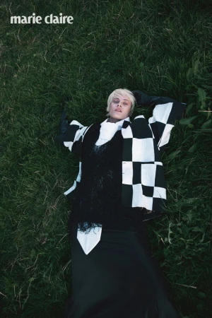 Model lying on deep green grass wearing a black and white checkered jacket with a high white collar and black gloves.