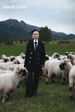 Model Paulina Heiler in a black double-breasted coat and tie standing amidst a flock of sheep in a mountainous meadow.