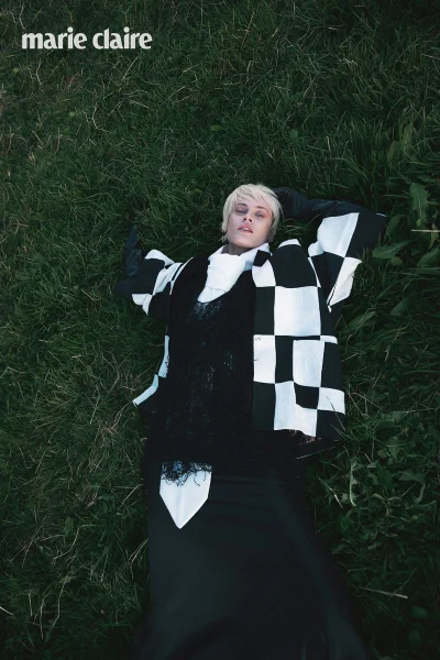 Model lying on deep green grass wearing a black and white checkered jacket with a high white collar and black gloves.